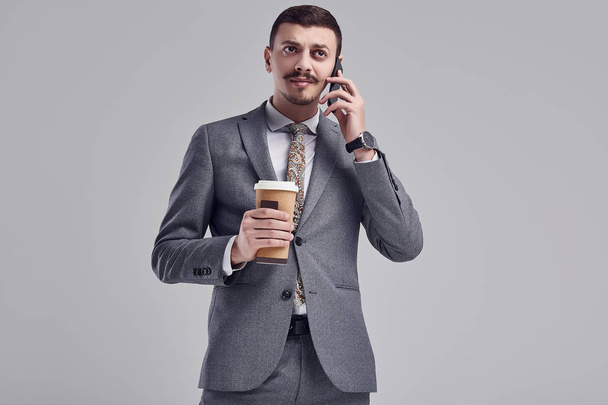 Portrait of handsome young confident arabic businessman with fancy mustache in fashion gray full suit holds a cup of coffee and talking on the phone - Zdjęcie, obraz