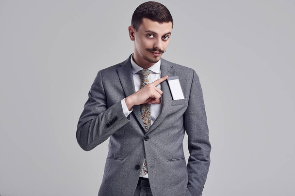 Retrato de belo jovem empresário árabe confiante com bigode fantasia em moda cinza terno completo escondendo o telefone em seu bolso no fundo do estúdio
 - Foto, Imagem