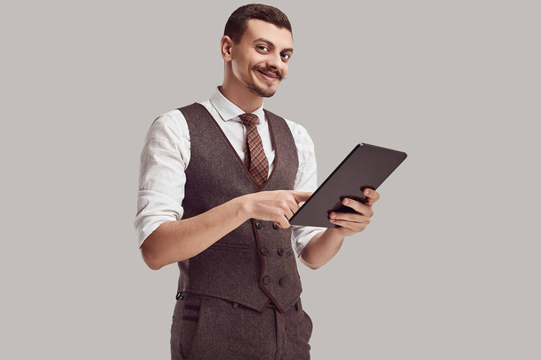 Portret van knappe jonge vertrouwen Arabisch zakenman met fancy snor in wollen bruin pak houdt tablet op studio achtergrond - Foto, afbeelding