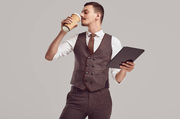 Portrait of handsome young confident arabic businessman with fancy mustache in woolen brown suit holds tablet and drink coffee on studio background - Photo, Image