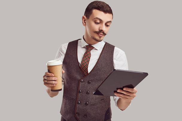 Csinos fiatal magabiztos Arab üzletember, díszes bajusz gyapjú barna öltönyt portréja tart tabletta és csésze kávé stúdió háttere - Fotó, kép