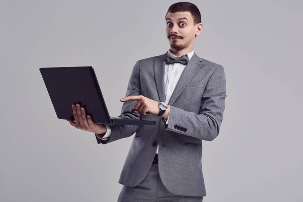 Porträt eines hübschen jungen selbstbewussten arabischen Geschäftsmannes mit schickem Schnurrbart im modischen grauen Ganzanzug hält schwarzen Laptop auf Studiohintergrund - Foto, Bild