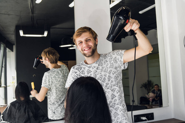Молодая женщина в парикмахерской. Красивый мужчина парикмахер сушит волосы с феном
 - Фото, изображение