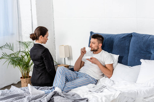 přítelkyně v obleku, sedící na posteli u přítele v pyžamu pití kávy v ráno ve všední den, koncept genderové stereotypy - Fotografie, Obrázek