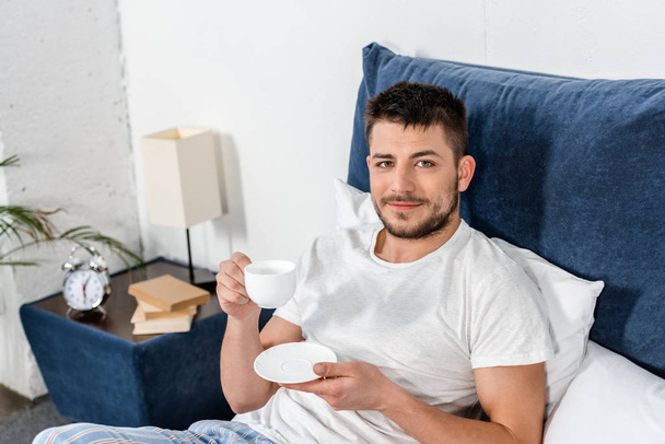 high angle view of handsome man holding cup of coffee in morning in bedroom and looking at camera - Fotó, kép