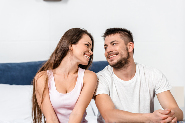smiling girlfriend and boyfriend in pajamas sitting on bed in bedroom and looking at each other - Photo, Image