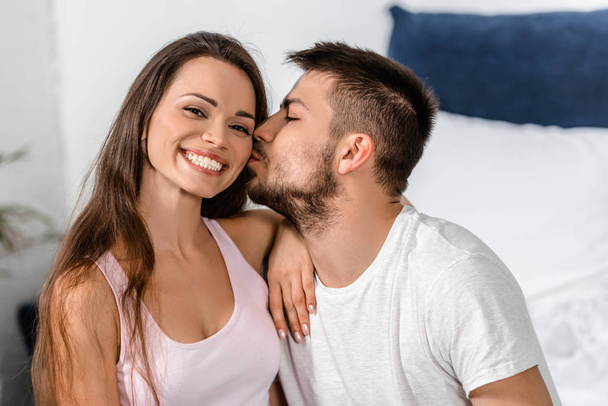 boyfriend in pajamas hugging and kissing smiling girlfriend on bed in bedroom - Photo, Image