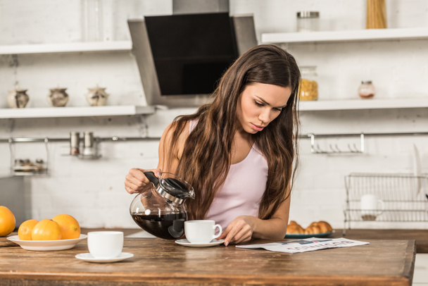 attractive woman reading business newspaper and pouring coffee into cup in morning at kitchen - Photo, Image
