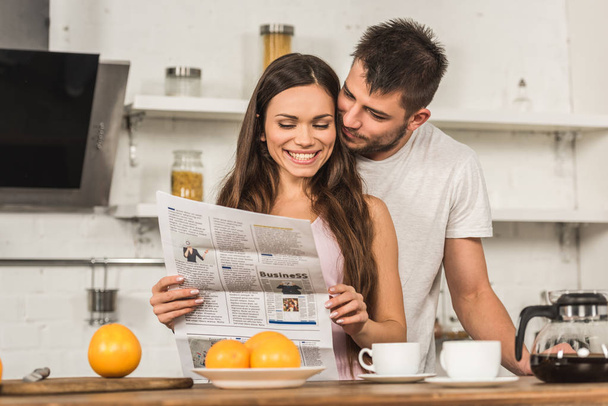 smiling girlfriend reading newspaper and boyfriend hugging her from back in morning at kitchen - Photo, Image