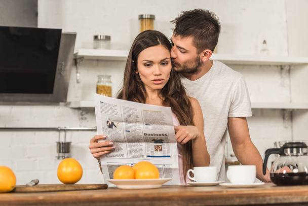 shocked girlfriend reading newspaper and boyfriend kissing her in morning at kitchen - Foto, imagen