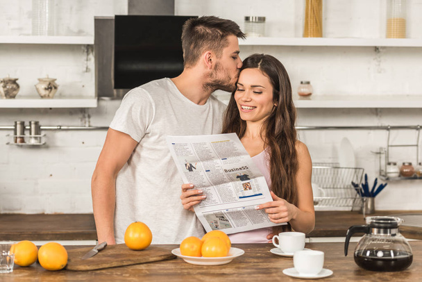 smiling girlfriend reading newspaper and boyfriend kissing her in morning at home - Фото, изображение
