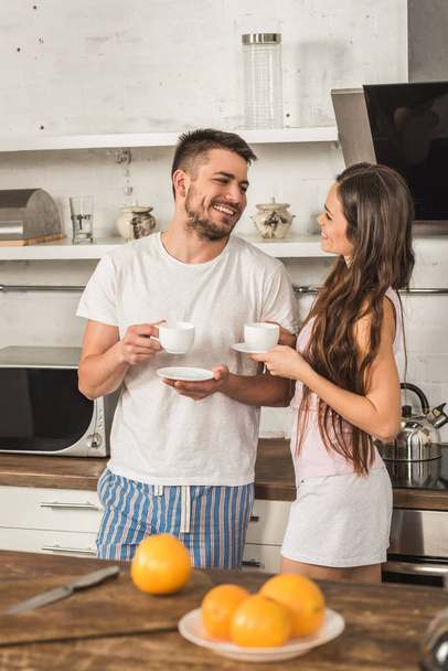 smiling girlfriend and boyfriend holding cups of coffee and looking at each other in morning at kitchen - Фото, зображення