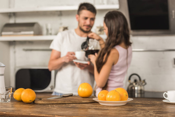 Selektivní fokus přítelkyně nalití kávy v šálku přítel, pomeranče na stole v kuchyni - Fotografie, Obrázek