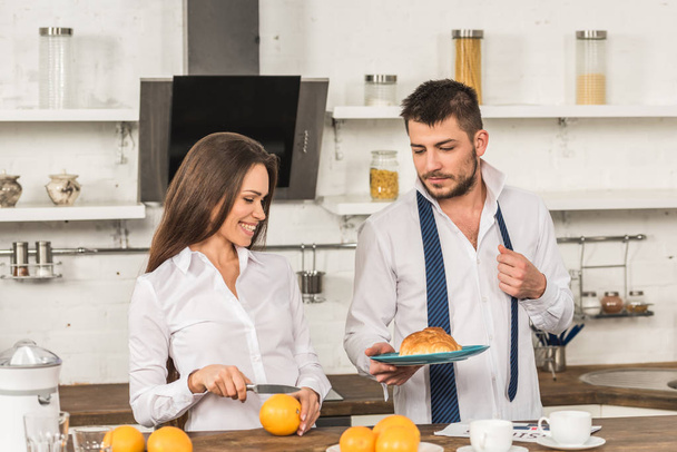 boyfriend holding plate with croissant and girlfriend cutting oranges at home, gender equality concept - Photo, Image