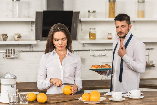 boyfriend holding plate with croissants and girlfriend cutting oranges at home - Photo, Image