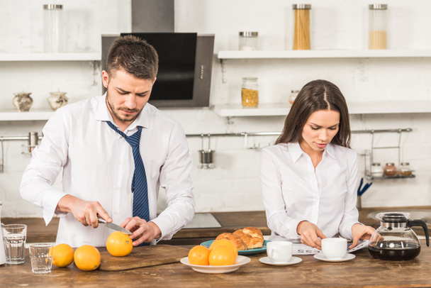 boyfriend cutting oranges and girlfriend reading newspaper at home, gender equality concept - Photo, Image
