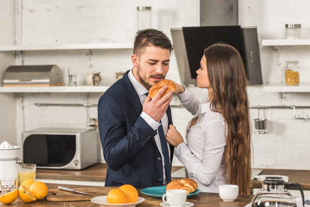 boyfriend eating croissant and girlfriend fixing his jacket in kitchen, sexism concept - Foto, Bild