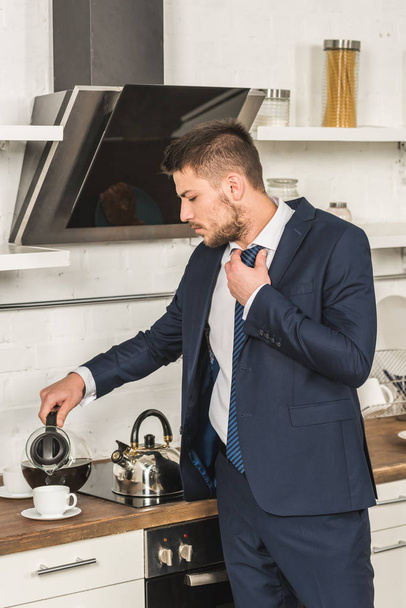 homem bonito em terno derramando café na xícara e amarrar gravata de manhã na cozinha
 - Foto, Imagem