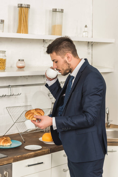Вид сбоку красивого мужчины в костюме, пьющего кофе и держащего круассан по утрам на кухне
 - Фото, изображение