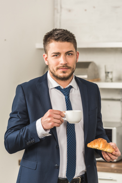 portrait of handsome man in suit holding cup of coffee and croissant and looking at camera in morning at kitchen - 写真・画像