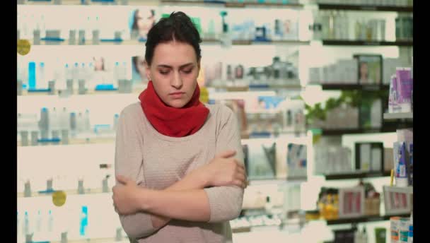 ein erkältetes Mädchen steht in einer Apotheke, sie hat Halsschmerzen, ihr Hals ist in ein rotes Tuch gehüllt - Filmmaterial, Video