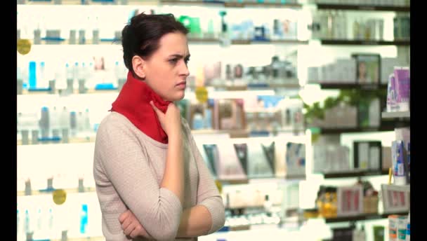 A cold girl stands in a pharmacy, she has a sore throat, her neck is wrapped in a red scarf - Footage, Video
