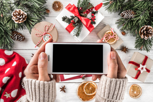 Mobiltelefon tartó női kezek. Karácsonyi ajándék dobozok, piros dekoráció, fenyő ágak, fenyőtobozok a holiday háttér. Karácsonyt és boldog új évet téma. Lapos feküdt, top view - Fotó, kép