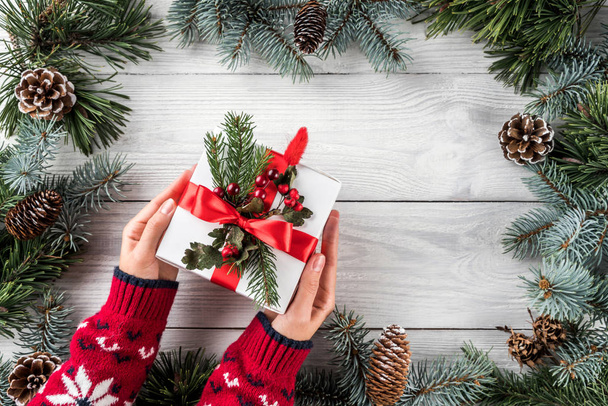 Руки женщины держат рождественскую подарочную коробку на белом деревянном фоне с еловыми ветками, сосновыми шишками, снежинками. Новогодняя тема. Плоский, вид сверху
 - Фото, изображение