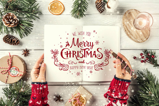 Female hand writing a letter to Santa. Christmas fir branches with holiday book, pencil, pine cones, gifts on white background. Xmas and Happy New Year theme. Flat lay, top view - Photo, Image