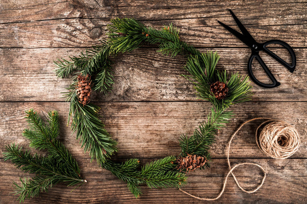 Рождественский венок из еловых ветвей, конусов, ножниц и джута на темном деревянном фоне. Новогодняя тема, снежинки. Плоский, вид сверху
 - Фото, изображение