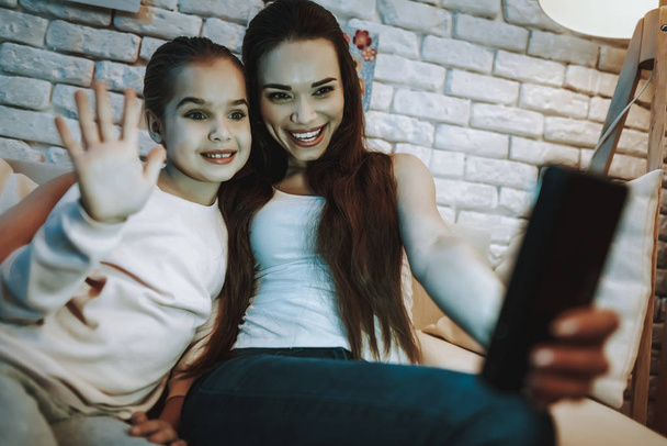 Mother with Daughter is Sitting on Couch. People Have a Video Call on Mobile Phone. Girl is Waving. Persons is Smiling. Wall with Pictures on Background. Evening Time. Home Interior. - 写真・画像