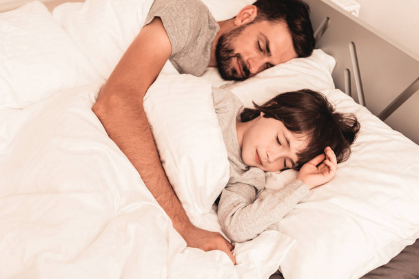 Little Son with Tired Sleeping Father in Bed. Family in Morning. Happy Family Concept. White Bed in Room. Father and Son at Home. Morning Routine. Lying in Bed. Sleeping Man. Blanket and Pillows. - Foto, afbeelding