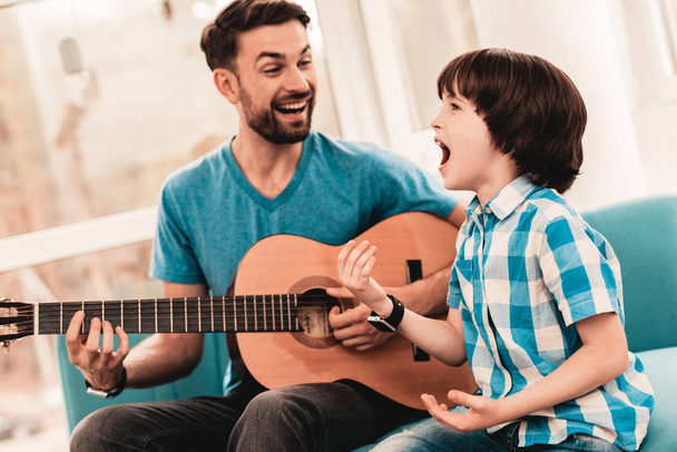 Young Bearded Father Playing on Guitar with Son. Happy Family Concept. Musician at Home. Young Boy in Shirt. Modern Hobby Concept. Music and Songs Concepts. Smiling Man. - 写真・画像