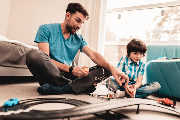 Bearded Father and Son Playing with Toy Race Road. Man Sitting on Floor. White Carpet in Room. Toy Cars. Exited Boy. Happy Family Concept. White Carpet. Lying on Floor. Indoor Fun. - Фото, изображение