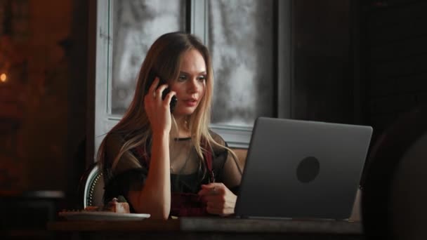Jovem mulher sentada no café na mesa de madeira, bebendo café e usando smartphone.On mesa é laptop. Menina navegando na internet, conversando, blogando. Feminino segurando telefone e olhando em sua tela
. - Filmagem, Vídeo
