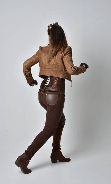retrato de longitud completa de chica morena con traje steampunk de cuero marrón. pose de pie con la espalda a la cámara, sobre fondo gris estudio
. - Foto, Imagen