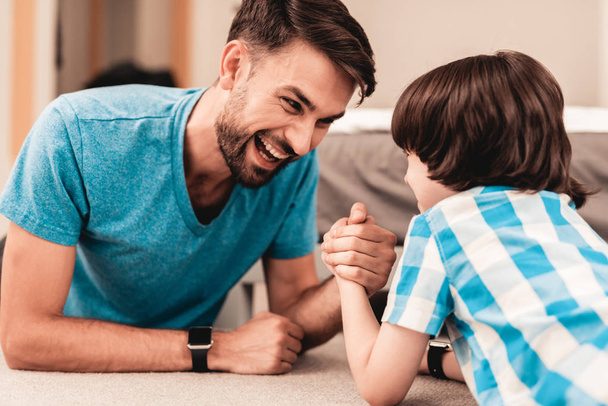 Father and Son Lying on Floor and Have Fun at Home. Sitting Boy. Young Father. Young Bearded Man at Home. Gray Sofa in Room. Happy Family Concept. White Carpet. Lying on Floor. Indoor Fun. - Foto, Imagen