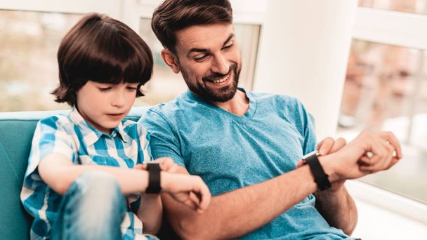Little Boy with Father Wacthing on Hand Watches. Sitting Boy. Young Father. Young Bearded Man at Home. Sitting on Blue Sofa. Happy Family Concept. Time Concepts. Modern Hand Watch. - Zdjęcie, obraz