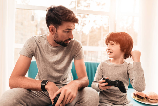 Father and Son Sitting and Playing on Console. Sitting Boy. Young Father. Playing on Console. Joystick in Hands. Young Bearded Man at Home. Sitting on Blue Sofa. Happy Family Concept. - Φωτογραφία, εικόνα
