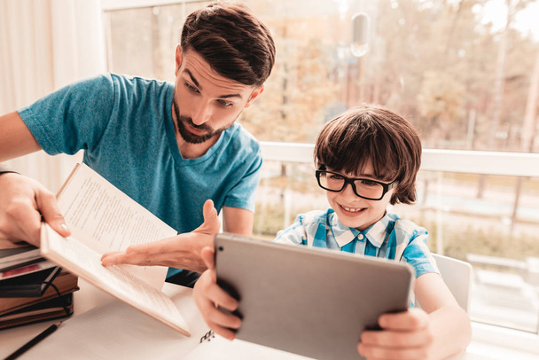 Father Trying to Help Son in Glasses with Homework. Education at Home. White Table in Room. Boy Using Digital Divece. Young Father. Lesson at Home. Education Concept. Boy in Shirt. Playing on Tablet. - Фото, изображение