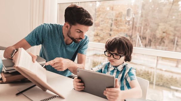 Father Trying to Help Son in Glasses with Homework. Education at Home. White Table in Room. Boy Using Digital Divece. Young Father. Lesson at Home. Education Concept. Boy in Shirt. Playing on Tablet. - Фото, зображення