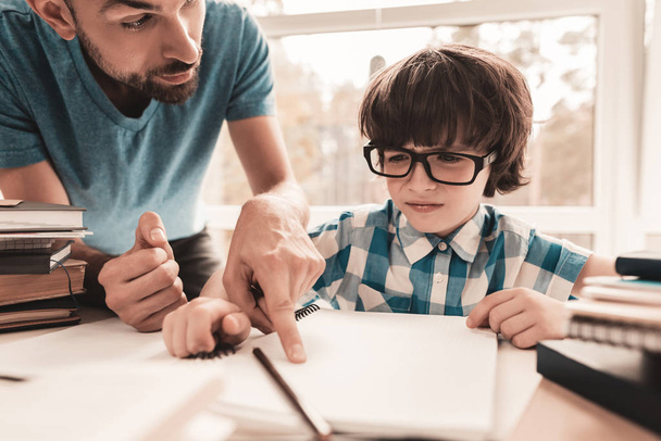 Little Boy in Glasses Doing Homework with Father. Education at Home. White Table in Room. Sitting Boy. Young Father. Lesson at Home. Education Concept. Writing in Notebook. Boy in Shirt. - Photo, Image