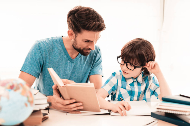 Little Boy in Glasses Doing Homework with Father. Education at Home. White Table in Room. Sitting Boy. Young Father. Lesson at Home. Education Concept. Writing in Notebook. Boy in Shirt. - Foto, Imagem