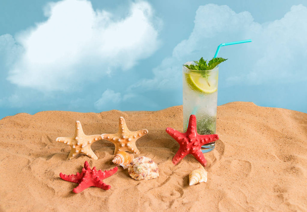 Glas Mojito-Cocktail, Seesterne und Muscheln am Sandstrand vor blauem Hintergrund. Sommerferienkonzept. - Foto, Bild