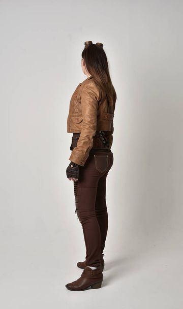 full length portrait of brunette  girl wearing brown leather steampunk outfit. standing pose with back to the camera, on grey studio background. - Photo, Image