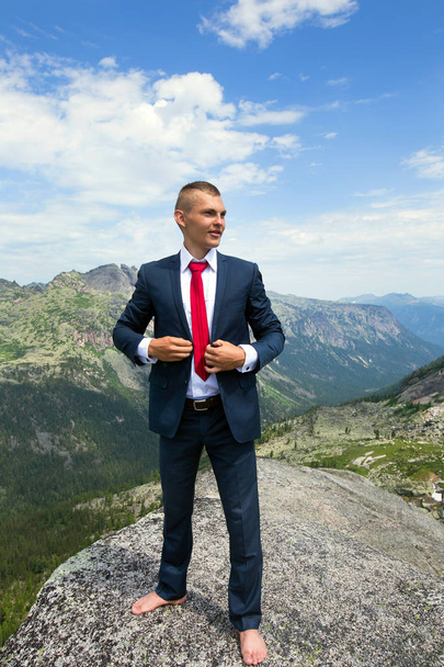 portrait of a young man in a business suit at the top of the mountain on the background of mountains and blue sky on a sunny summer day - Фото, изображение