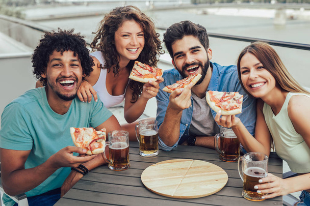 Друзі насолоджуються піцою. Група молодих веселих людей, які їдять піцу і п'ють пиво, сидячи в бобових мішках на даху будівлі
 - Фото, зображення