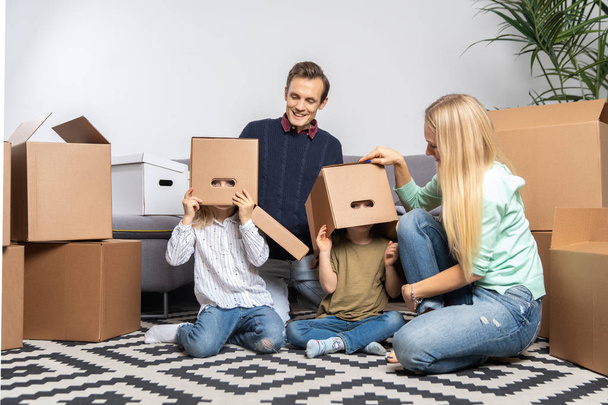 Kuva vanhemmista ja lapsista pahvilaatikot päässään istuu uudessa asunnossa
 - Valokuva, kuva