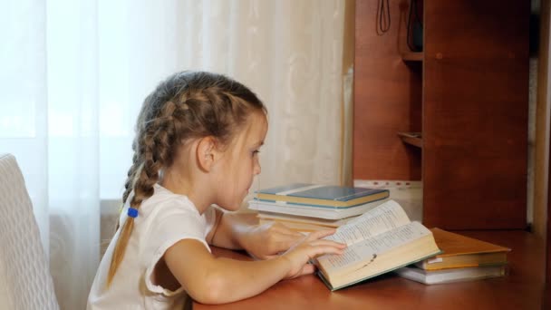 Widok z boku z małą dziewczynkę z plecionki siedzi w tabeli i czytania podręcznika odrabiania lekcji - Materiał filmowy, wideo
