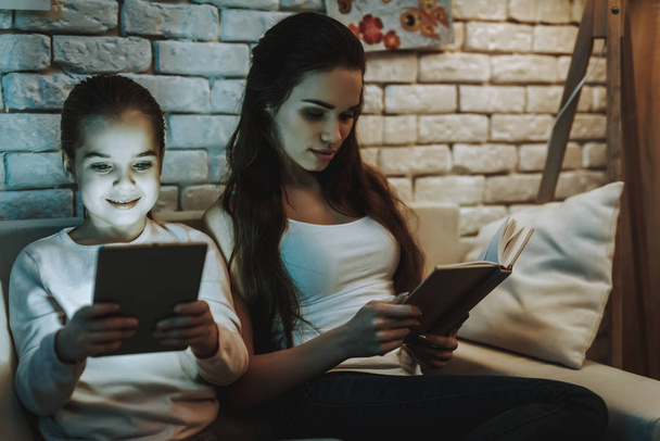 Mother with Daughter is Sitting on Couch. Mother is Reading a Book. Daughter is Watching Something on Tablet PC. Girl is Smiling. Wall with Pictures on Background. Evening Time. Home Interior. - Photo, image
